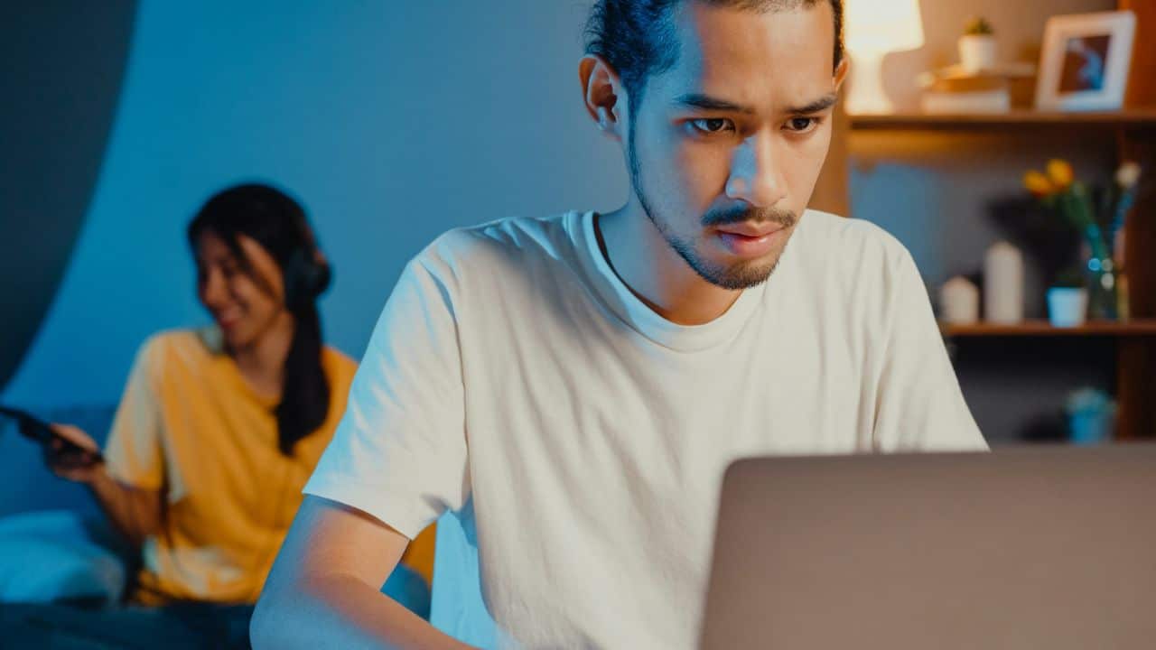 A man looks intently at a laptop while a woman smiles in the background.