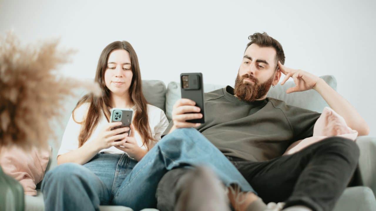 A couple sits on a couch, both looking at their phones.