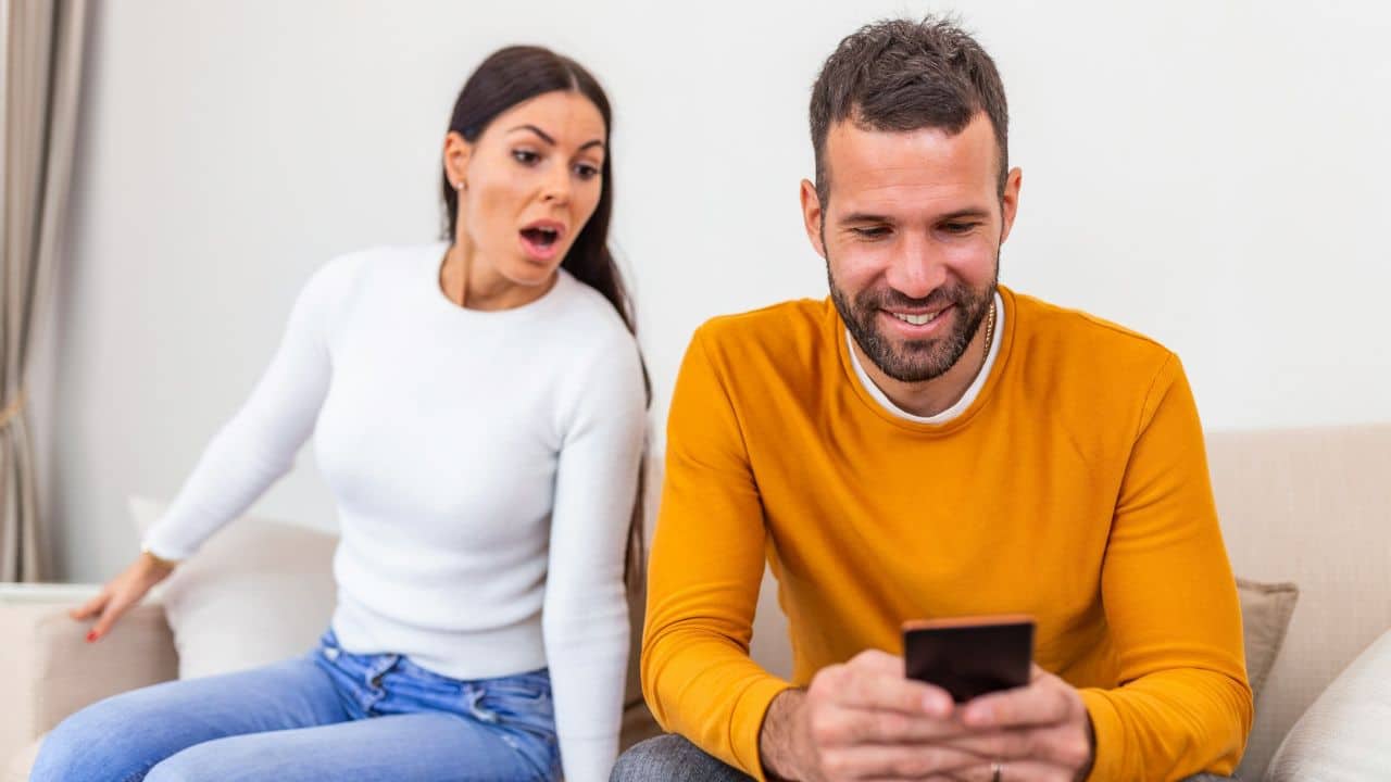 A smiling man looks at his phone while a surprised woman looks over his shoulder.