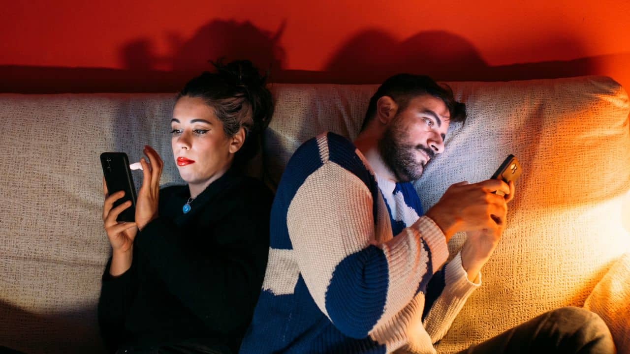 A couple sits back to back on a couch, both looking at their phones.