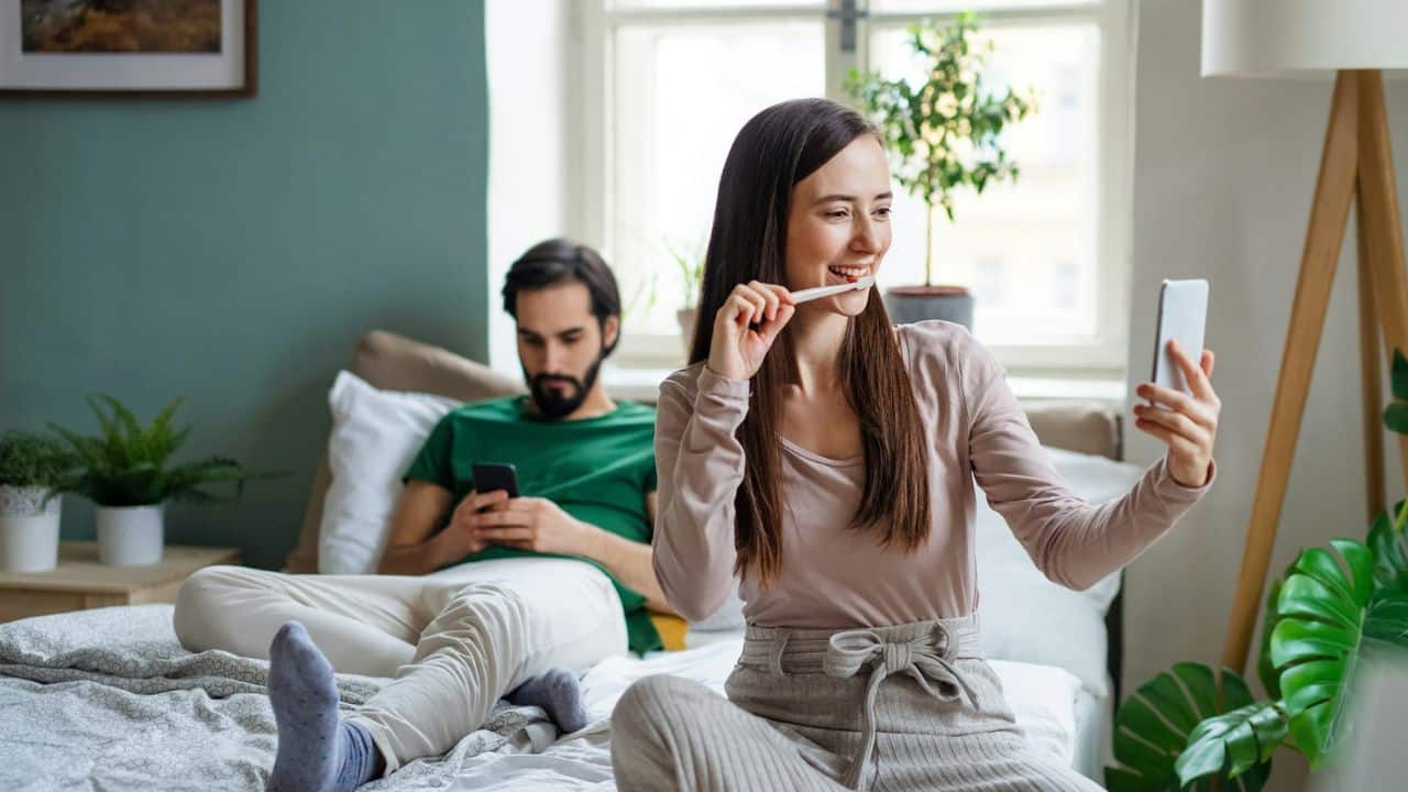 A woman smiles while taking a selfie on the bed as a man looks at his phone.