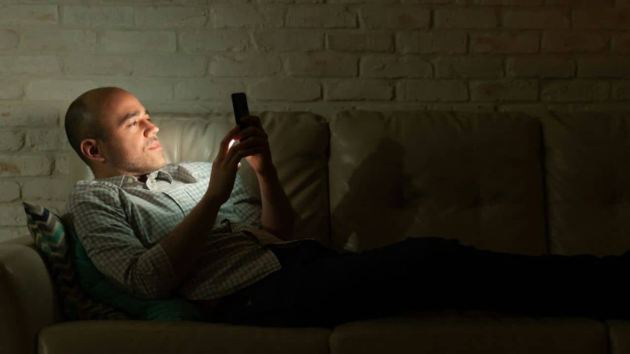A man lies on a couch in the dark, illuminated by his phone's light.