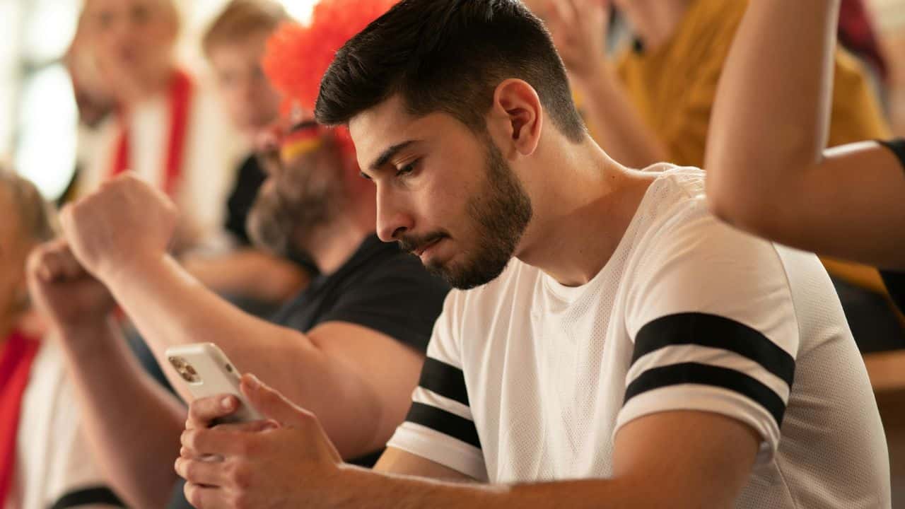 A man looks at his phone while a crowd celebrates in the background.