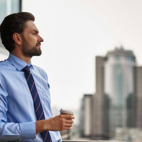 A man in a shirt and tie stands on a balcony looking at a city skyline.
