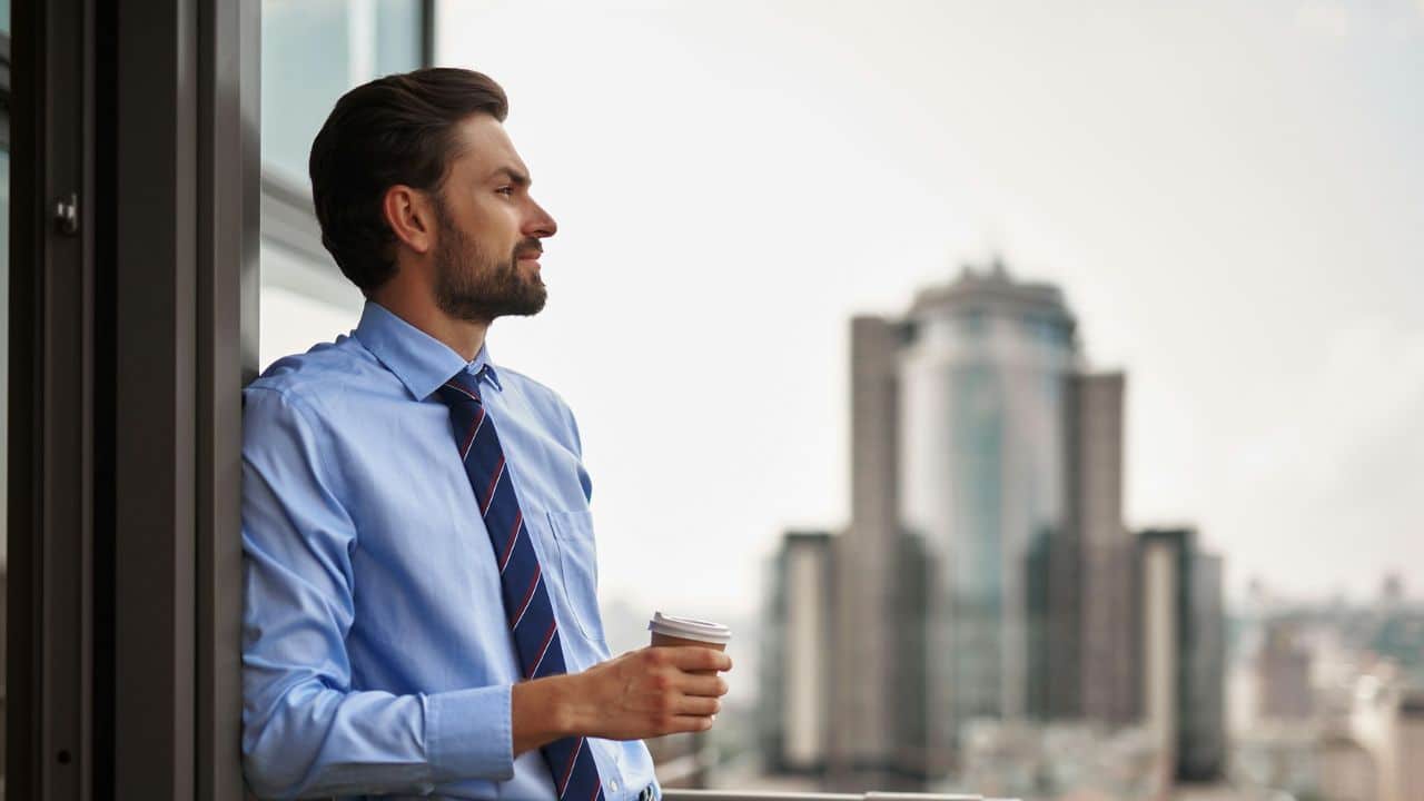 A man in a shirt and tie stands on a balcony looking at a city skyline.