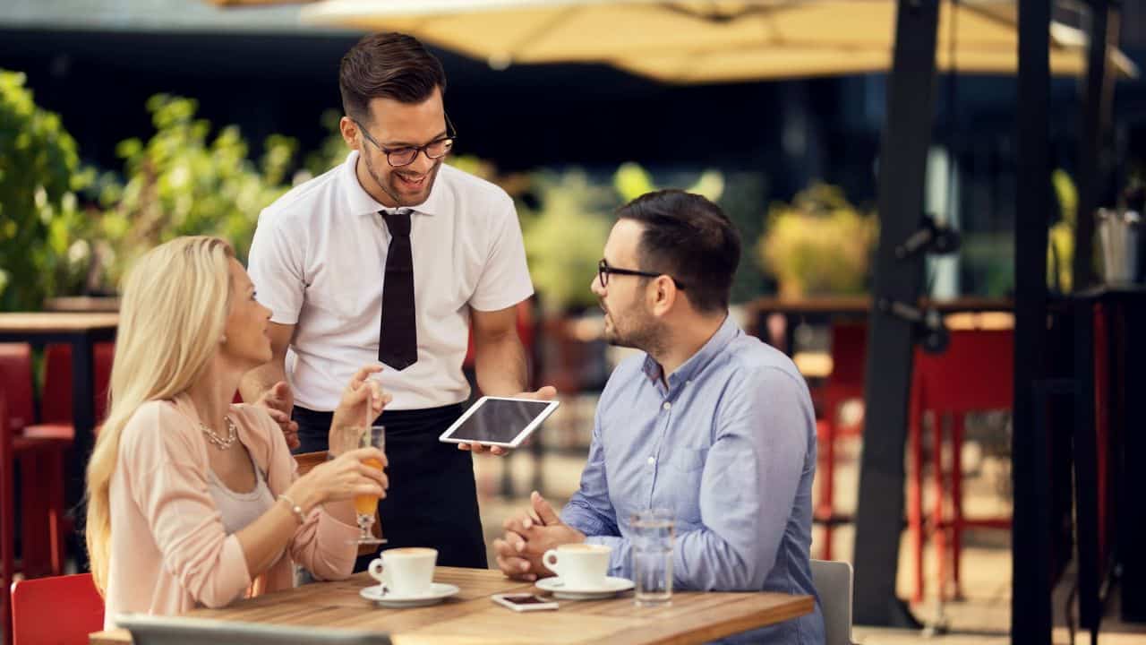 A waiter shows a tablet to a smiling couple at an outdoor cafe.