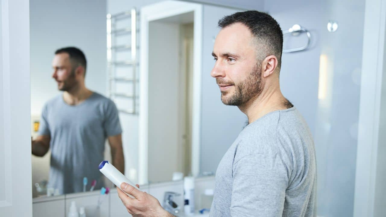 A man looks at his reflection in a bathroom mirror while holding deodorant.