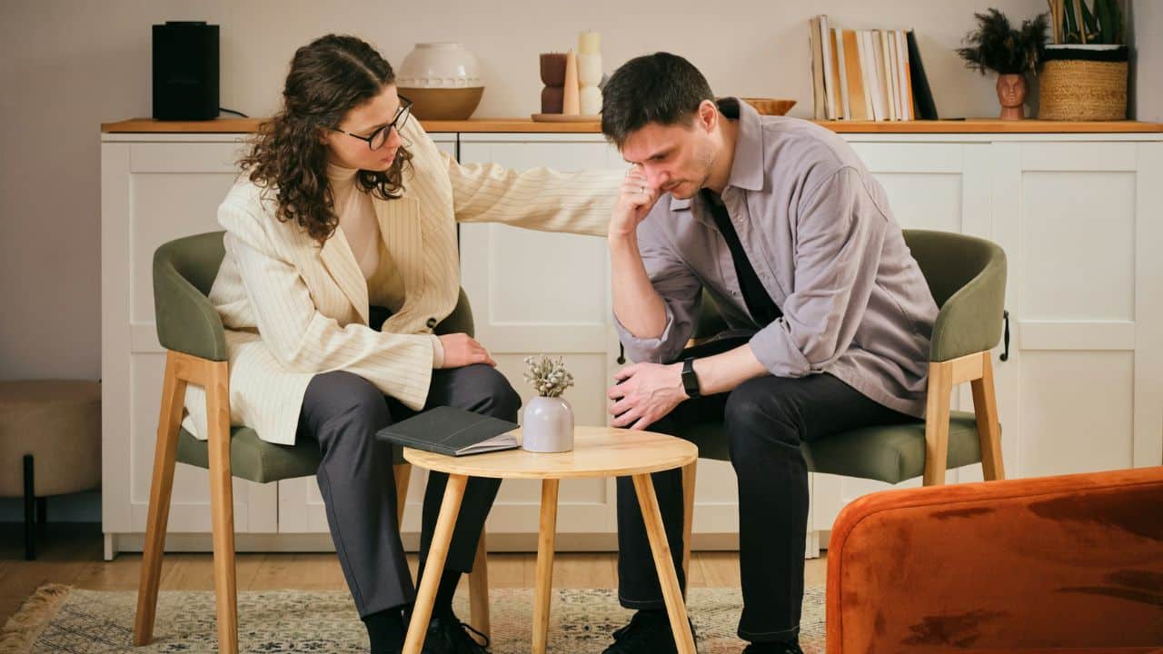 A woman comforts a sad man sitting in a chair with his head in his hand.