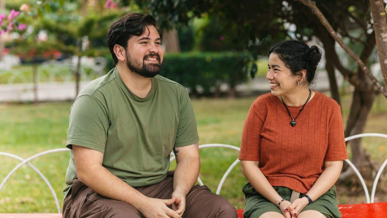 A happy couple is sitting on a bench in a park, smiling at each other.