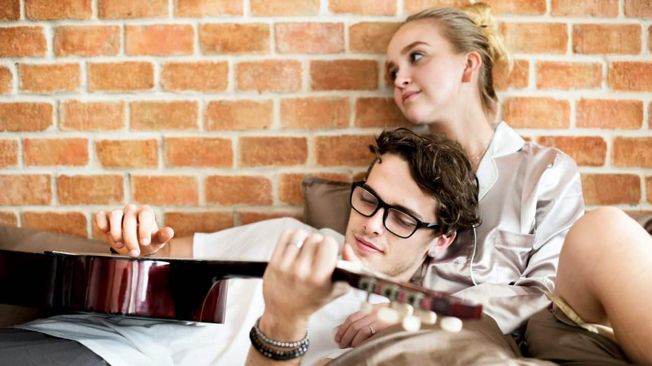A woman listens as a man plays an acoustic guitar on a brick wall.