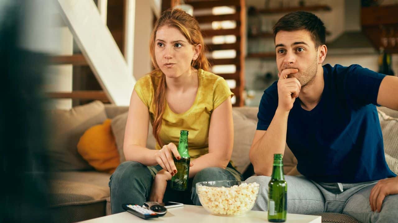 A couple sits on a couch watching TV with a bowl of popcorn and beers.