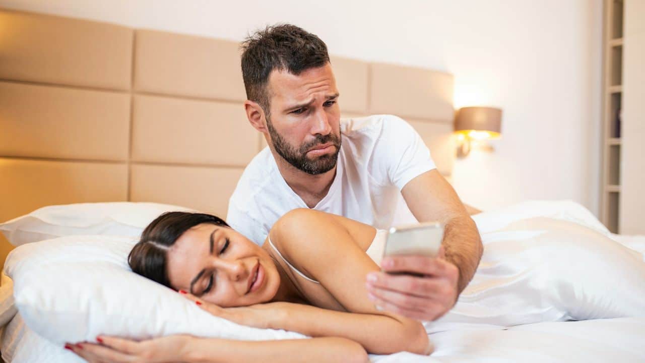 A man looks at a woman's phone while she sleeps next to him.