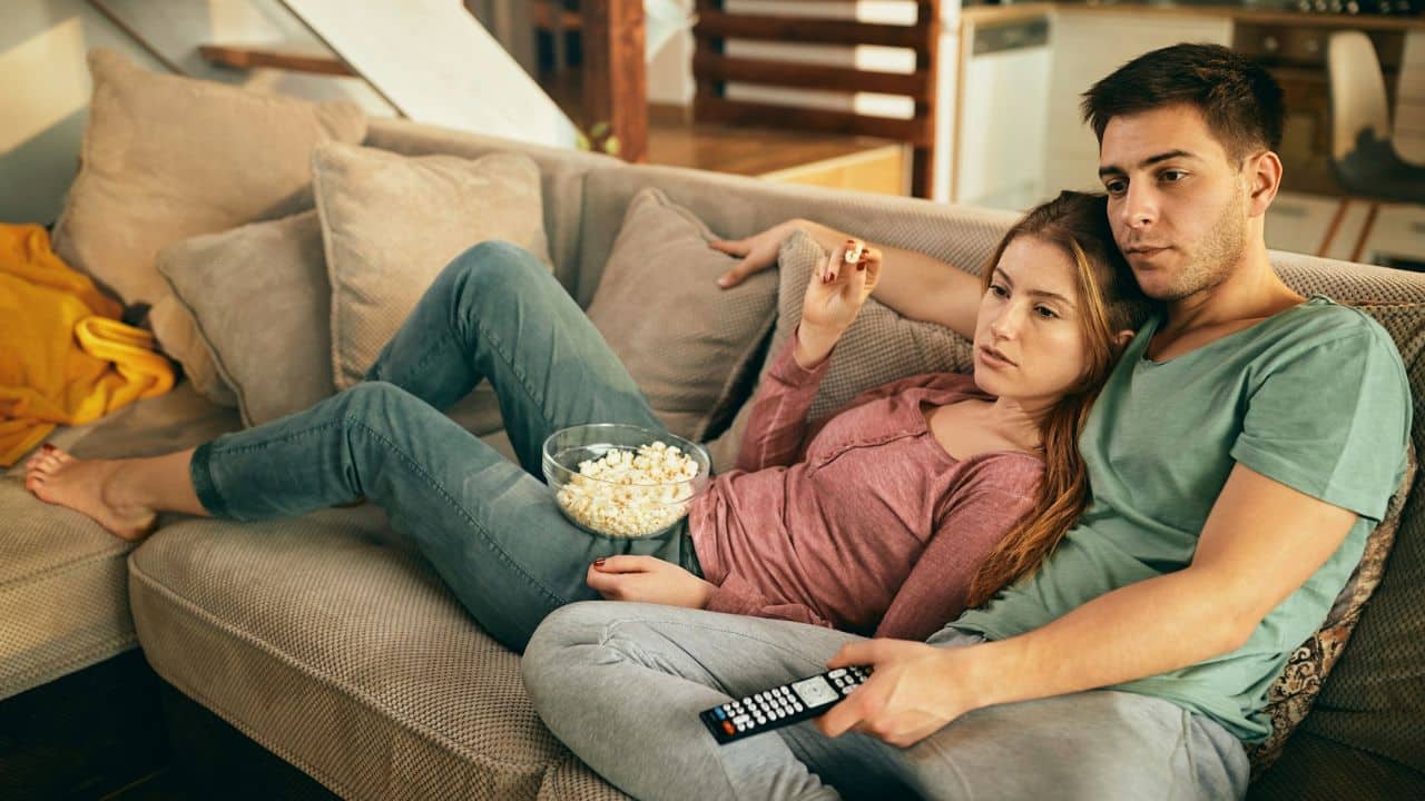 A couple reclines on a couch watching TV and eating popcorn.