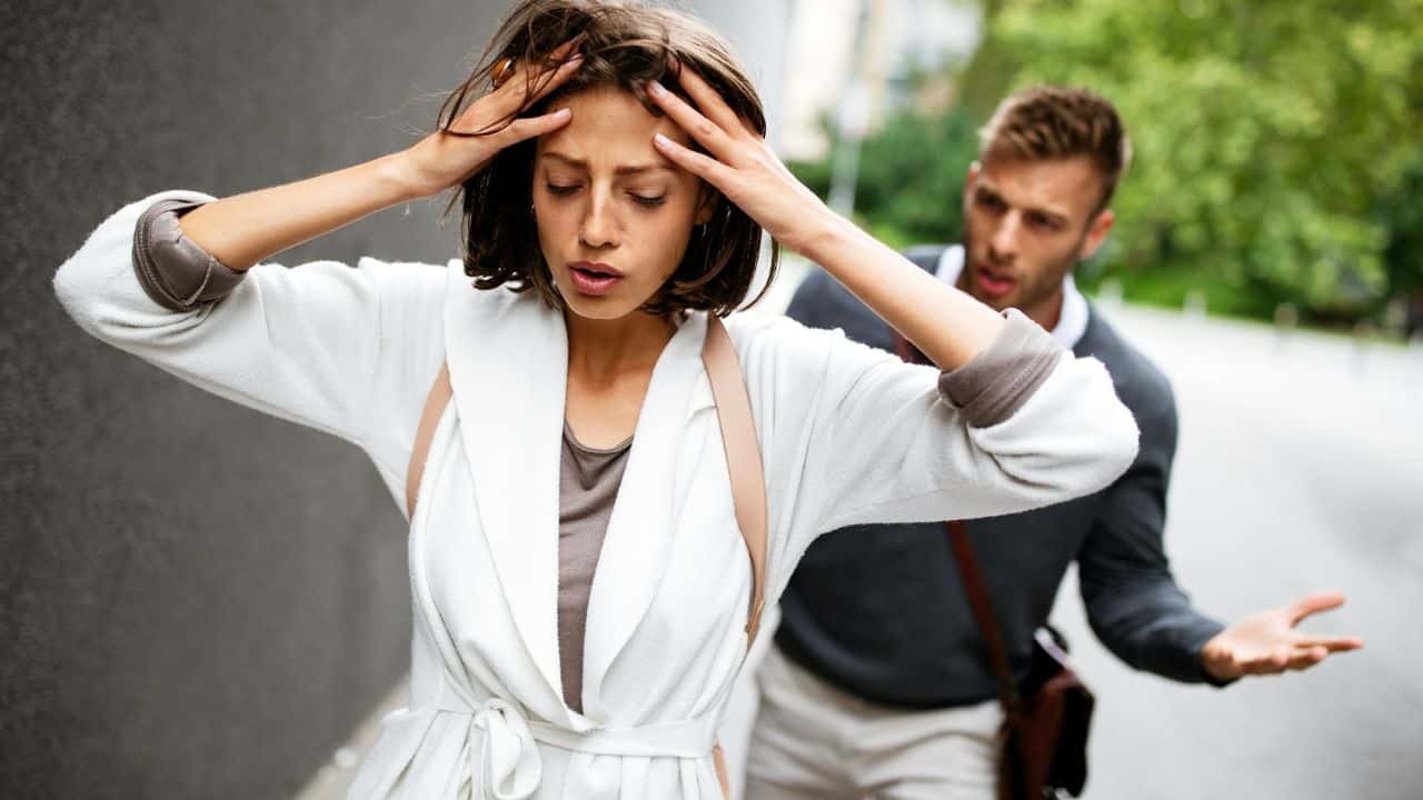 A woman looks distressed, holding her head as a man argues with her.