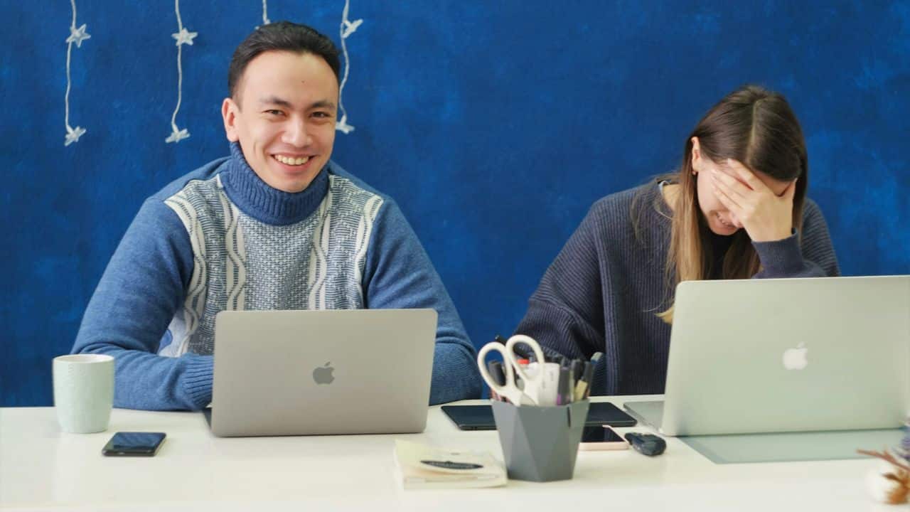 A man smiles at the camera as a woman next to him holds her hand to her face.