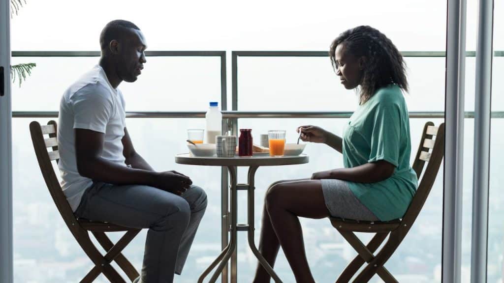 A serious-looking couple sits silently at a small table on a balcony.