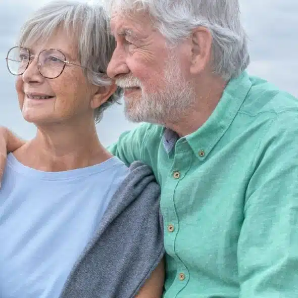 A smiling elderly couple embraces while looking out at the ocean.