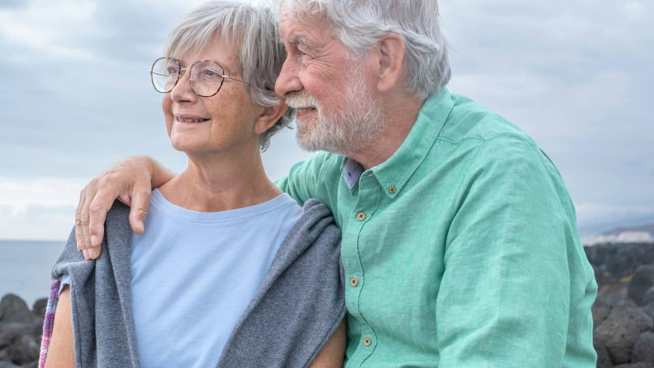 A smiling elderly couple embraces while looking out at the ocean.