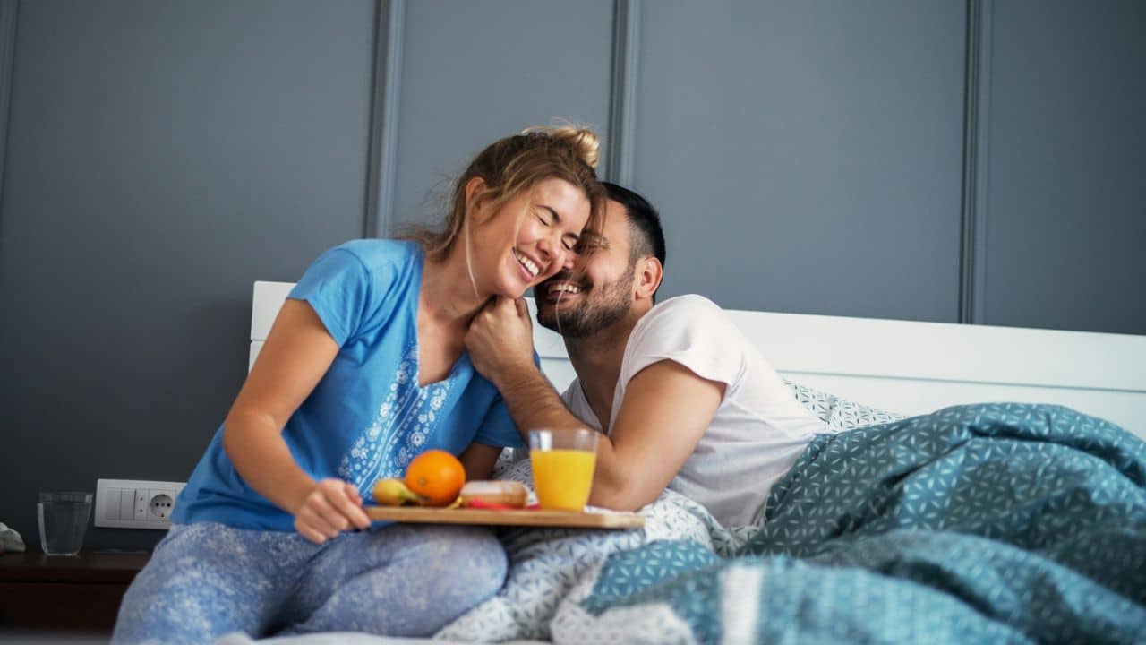A laughing couple enjoys breakfast in bed, with a tray of food and orange juice.