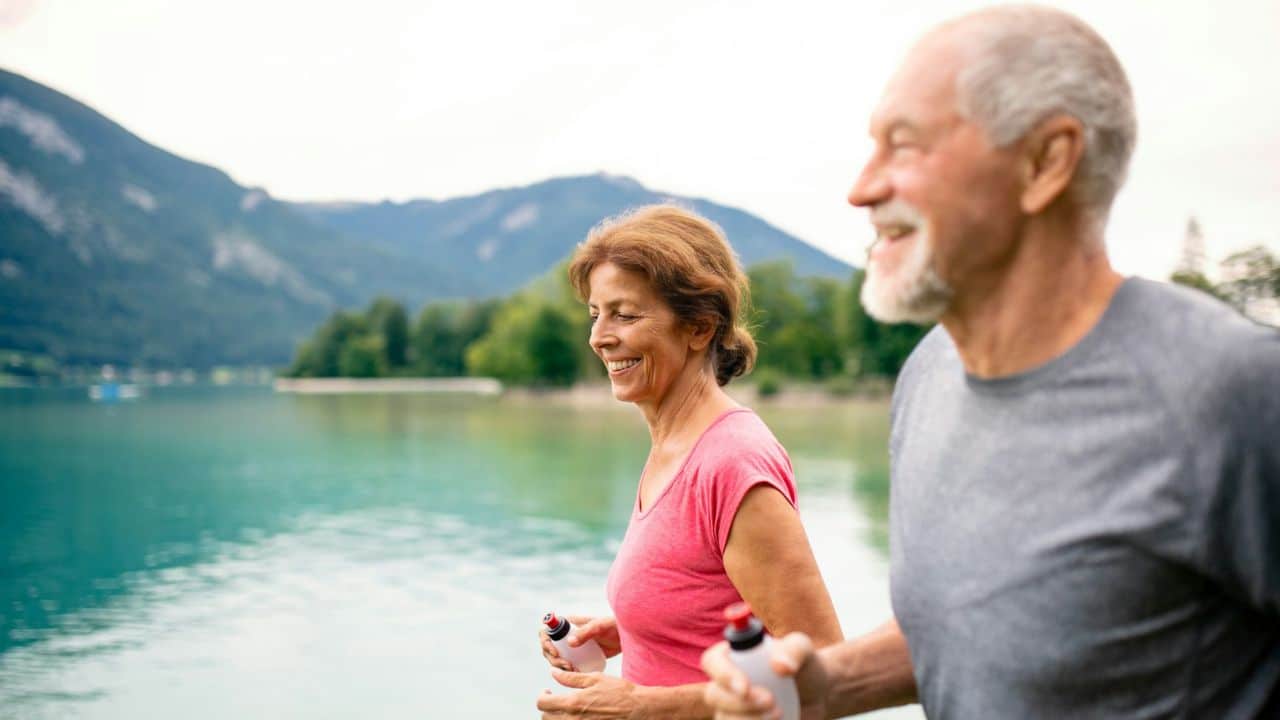 A happy older couple jogs together by a lake with mountains in the background.
