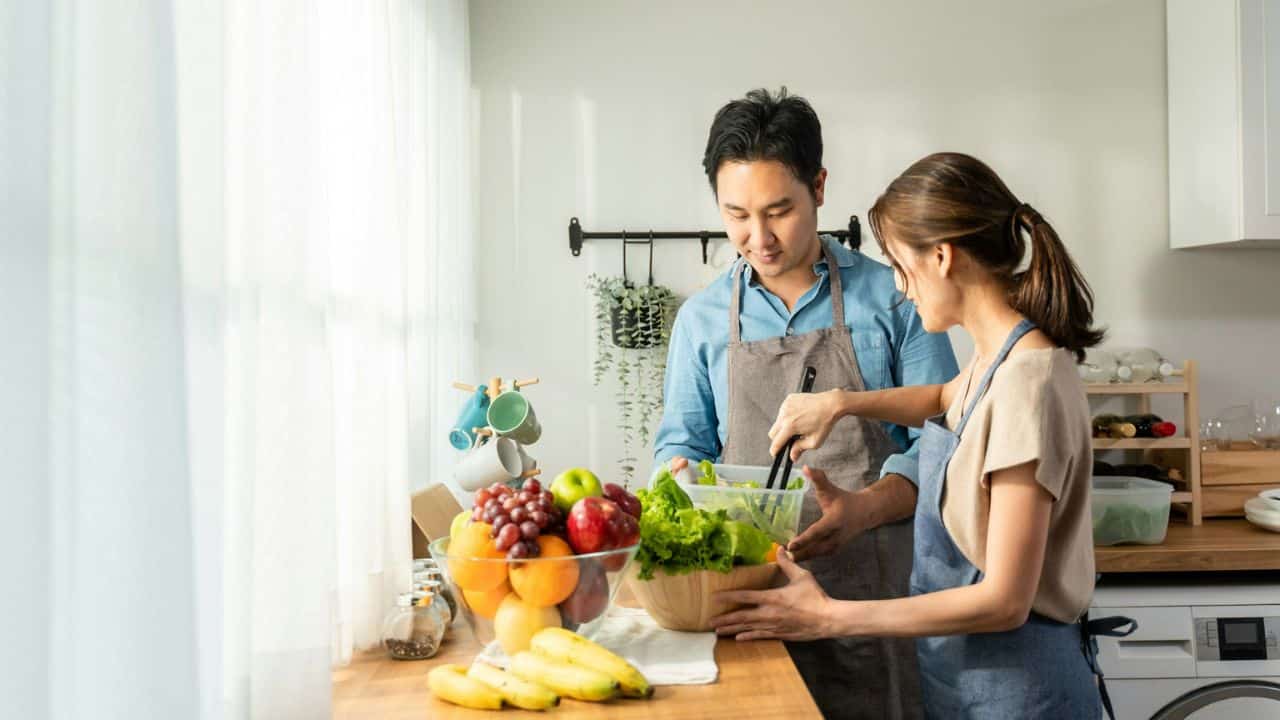 A smiling couple wearing aprons mixes a salad in a bright kitchen.