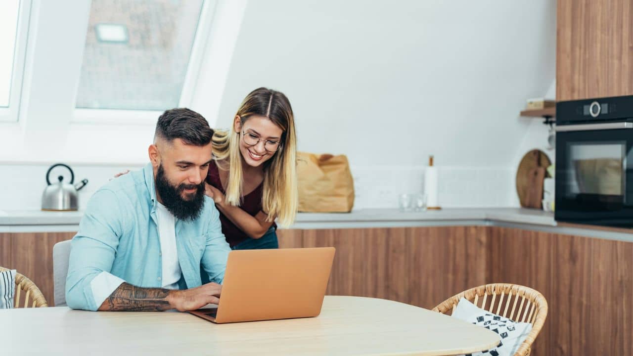 A couple sits at a kitchen table, looking at a laptop together.