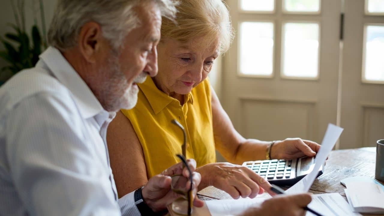 An elderly couple reviews documents and uses a calculator at a table.