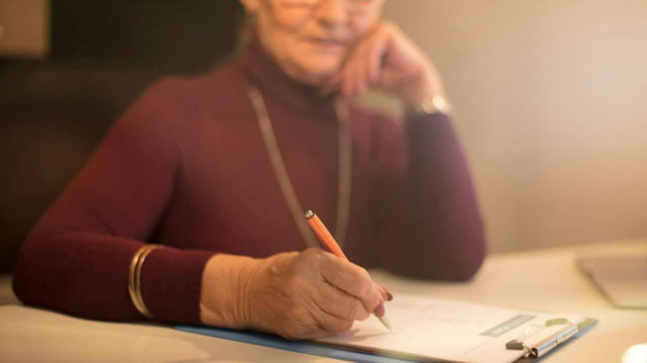 An older woman with glasses sits at a desk, writing on a document.