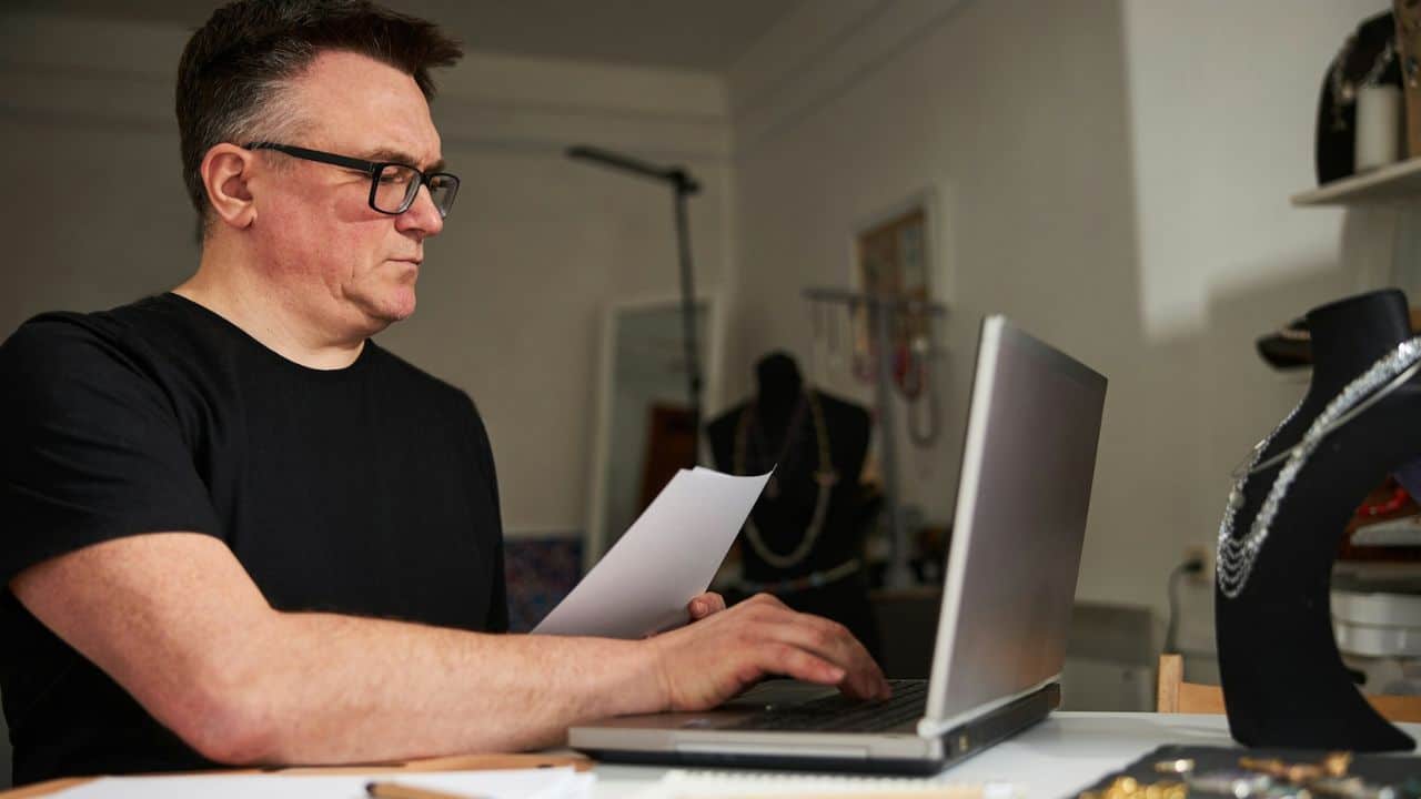 A bespectacled man in a black shirt works on a laptop while holding a document.