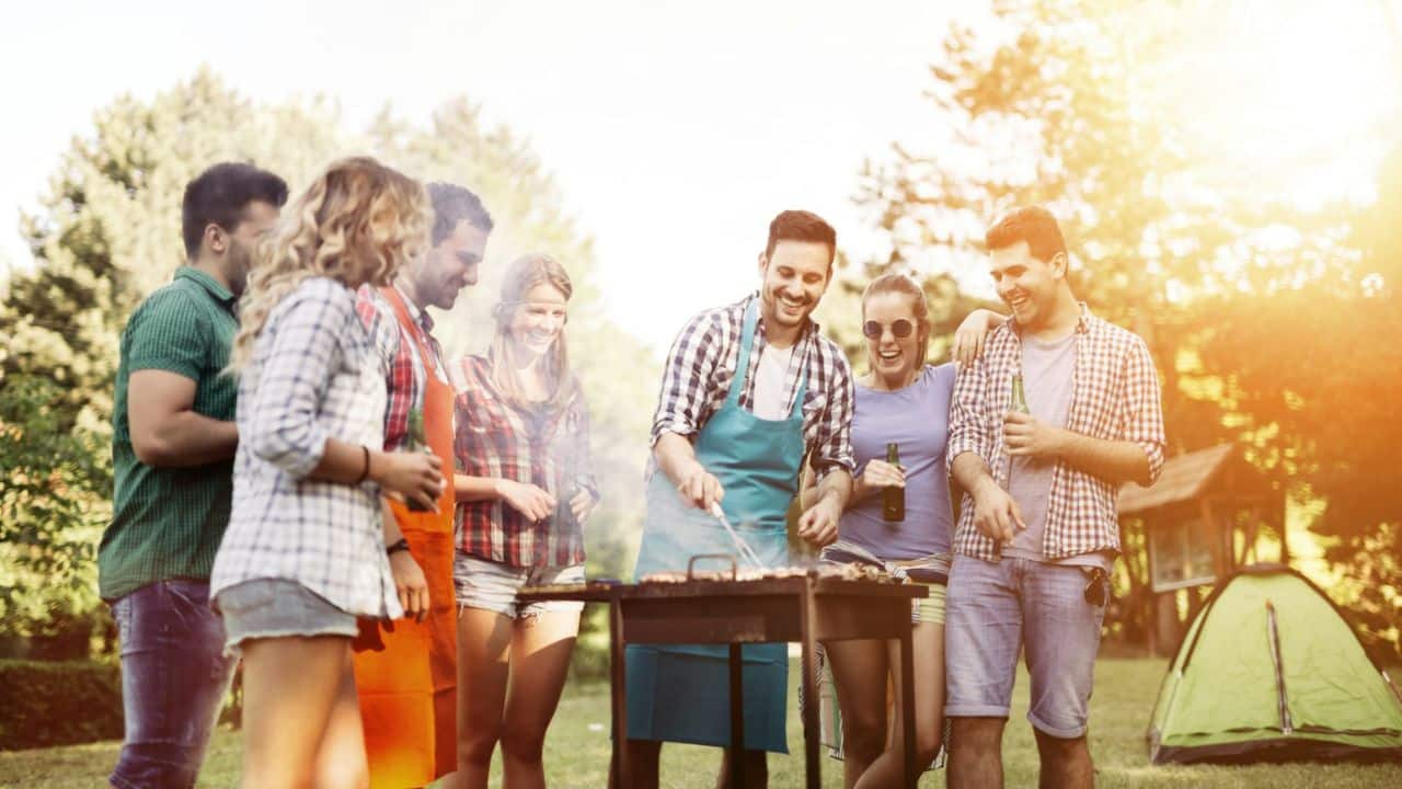A group of young adults laughs while a man grills food at a backyard barbecue.