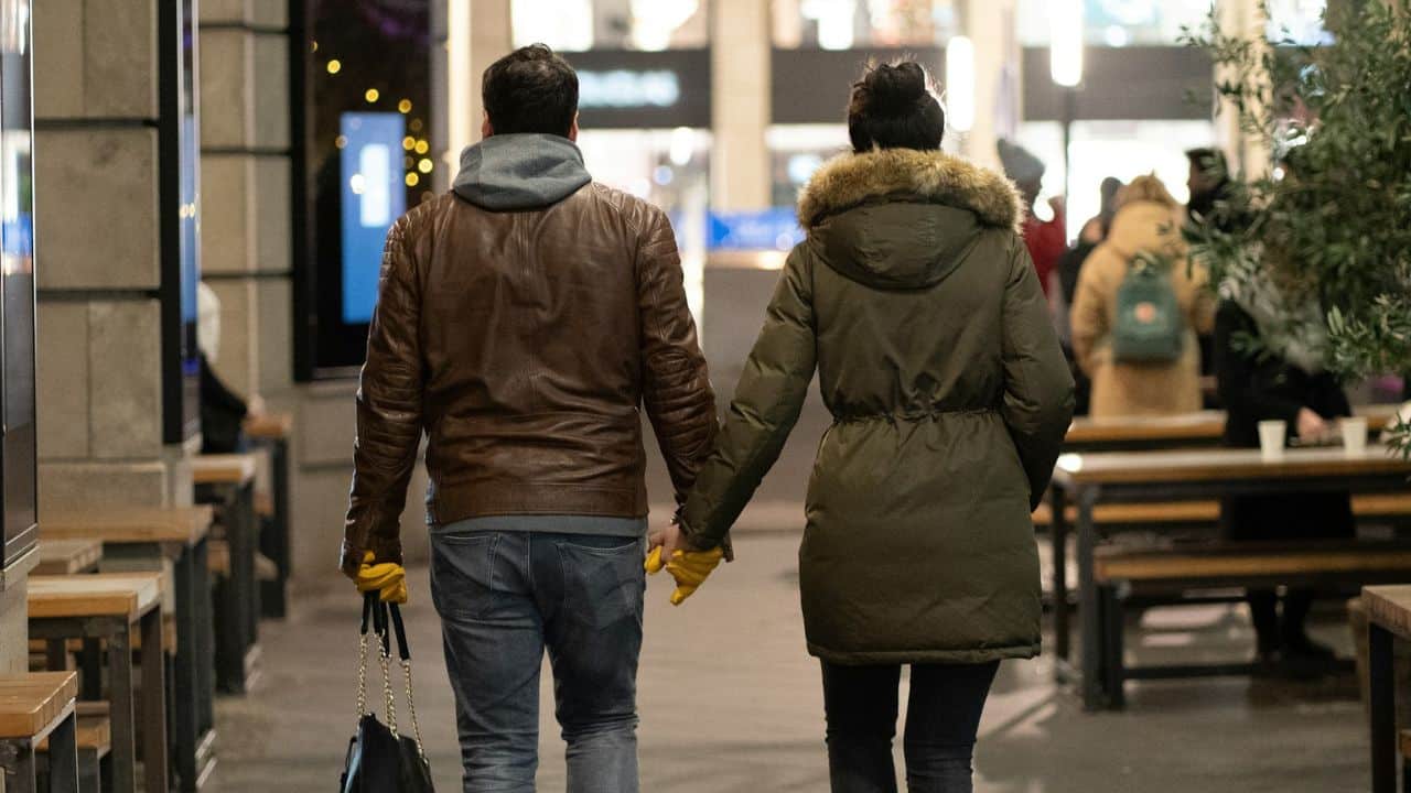 A couple holds hands from behind while walking down a street at night.