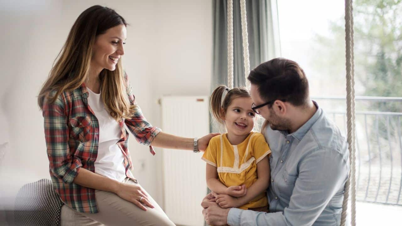 A happy family of three smiles as a father holds his daughter on a swing.