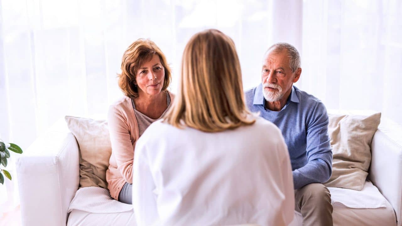 An older couple sits on a couch, facing a person in a white shirt.