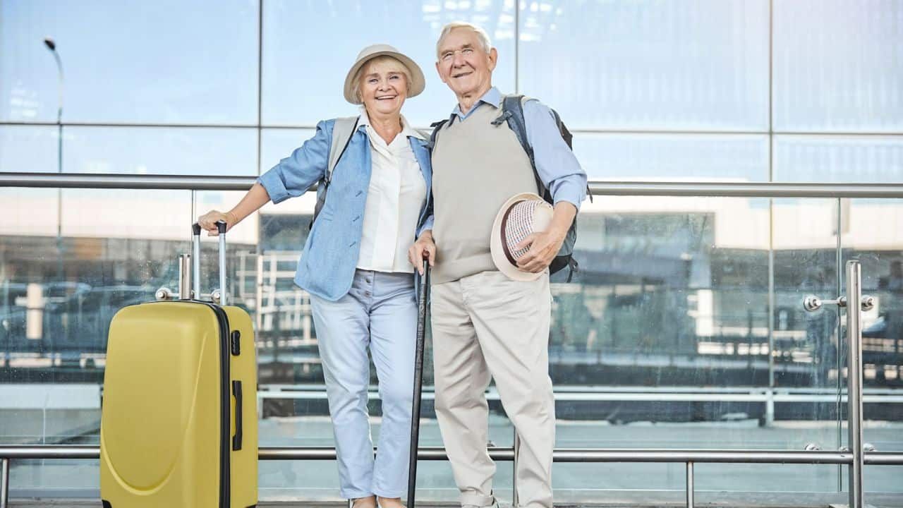A happy, smiling older couple stands at an airport with a yellow suitcase.