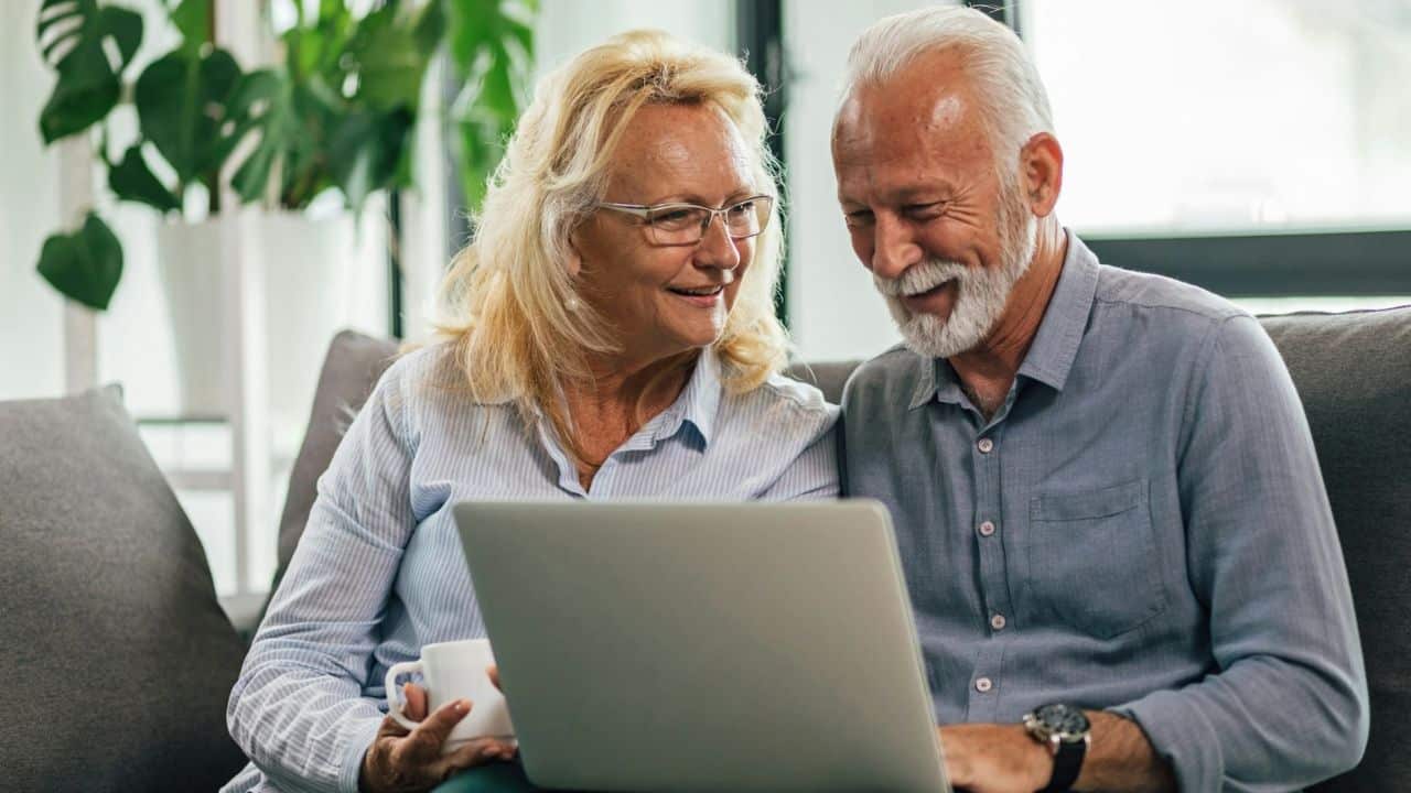 A happy, older couple sits on a couch, looking at a laptop together.