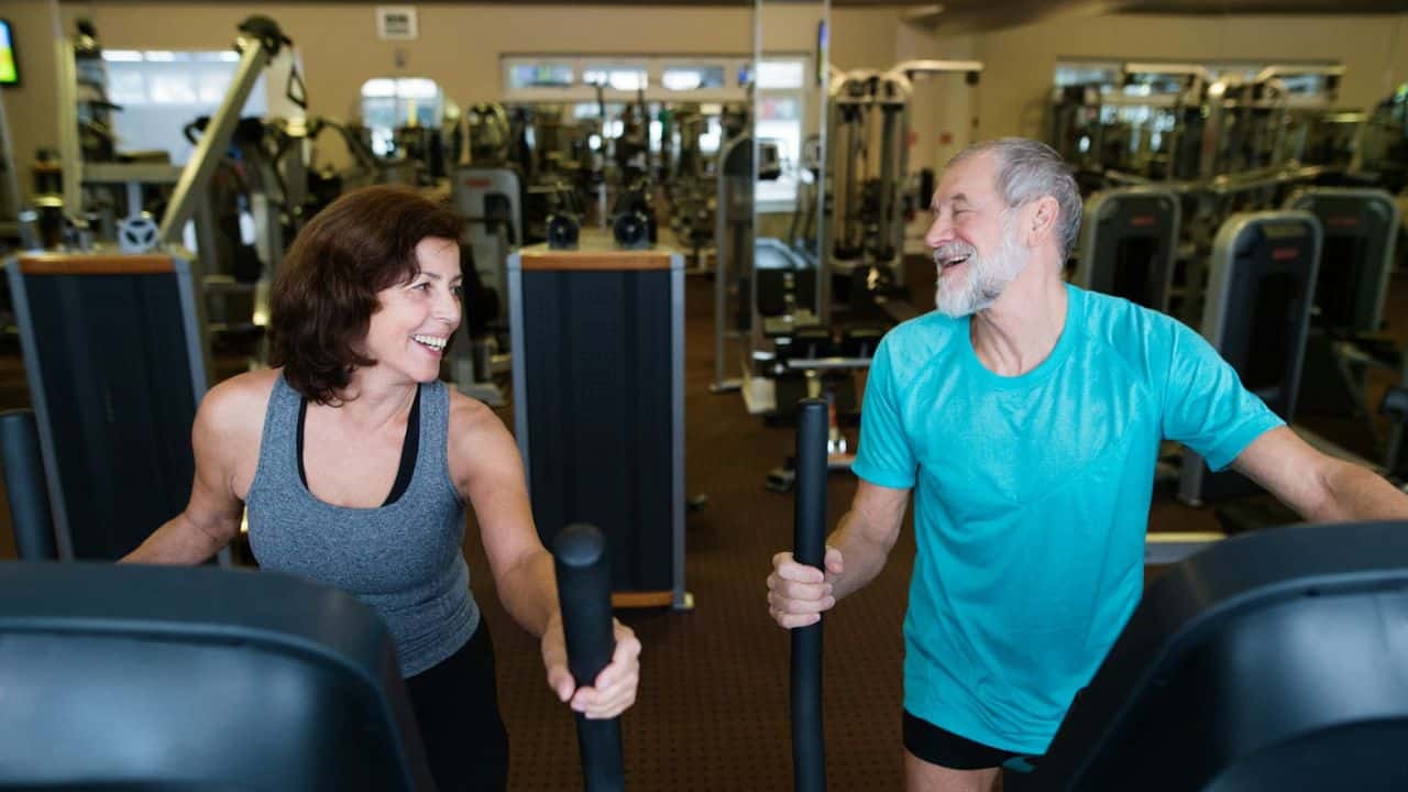 A laughing older couple works out together on exercise machines at a gym.
