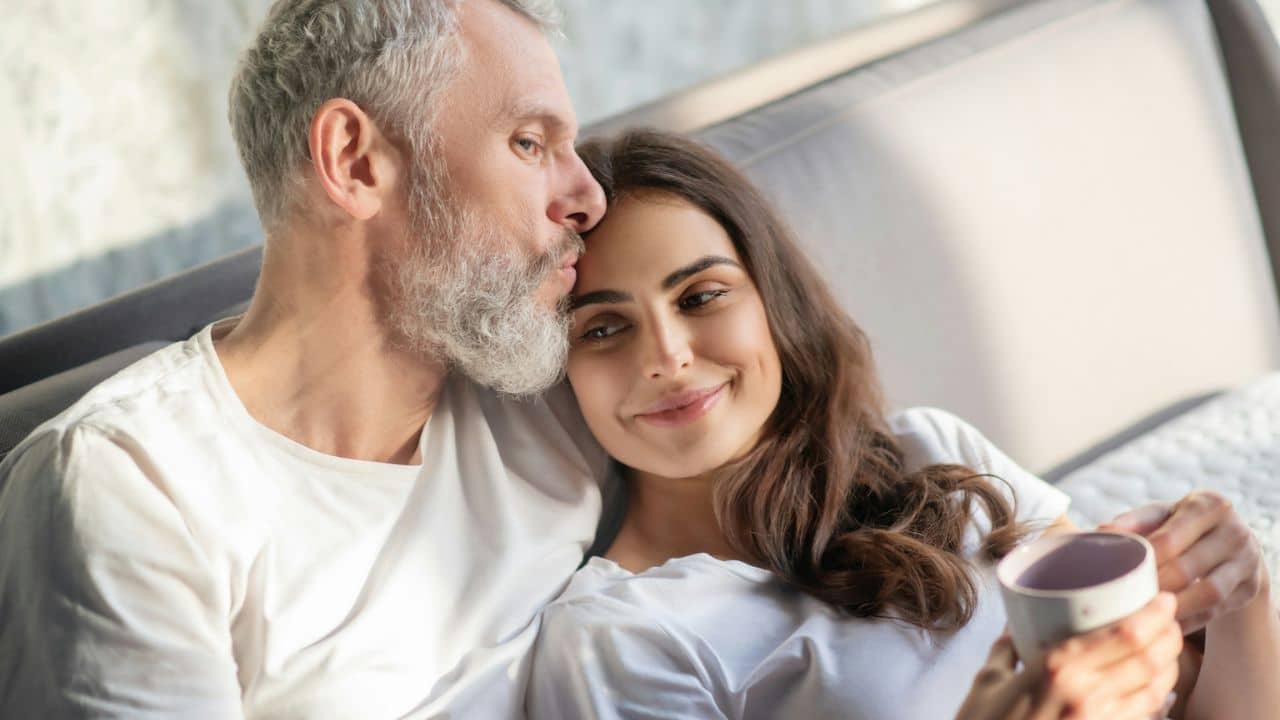 A bearded man in a white shirt kisses a smiling woman's forehead.