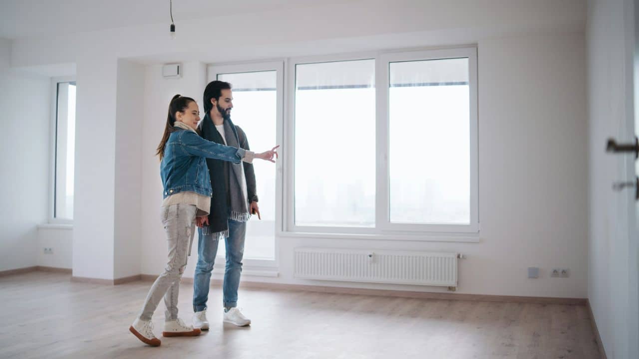 A young couple stands in an empty, bright room with large windows, looking and pointing.