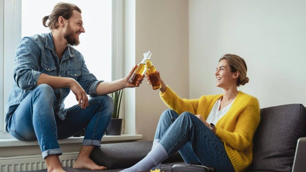 A smiling couple clinks beer bottles together in a sunlit living room.