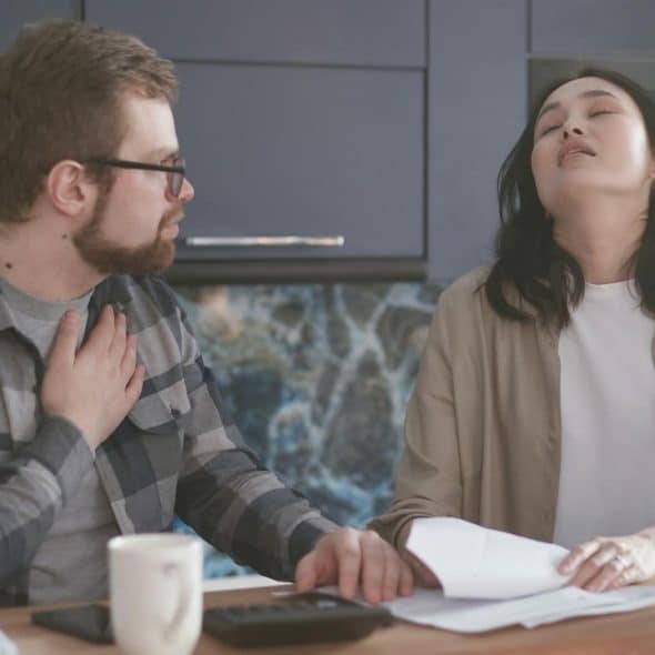 A frustrated woman with her eyes closed sits at a table while a man looks at her, holding his chest.