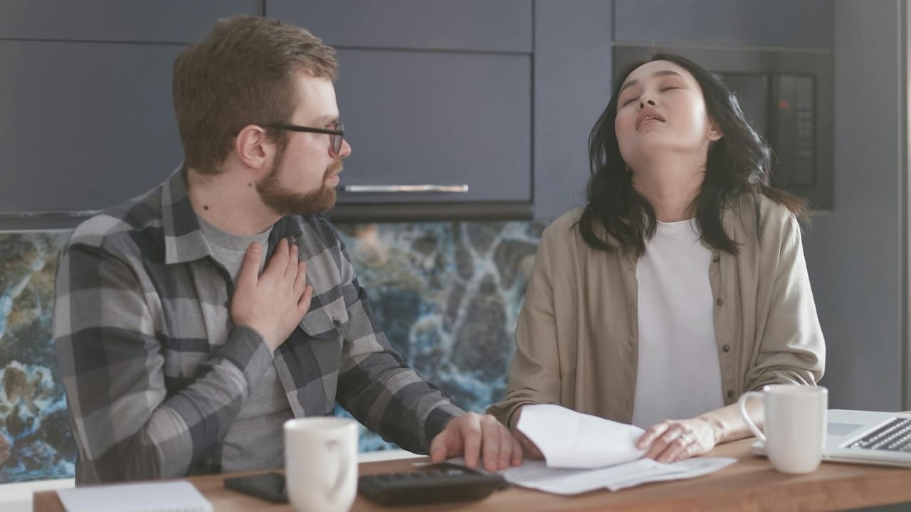 A frustrated woman with her eyes closed sits at a table while a man looks at her, holding his chest.
