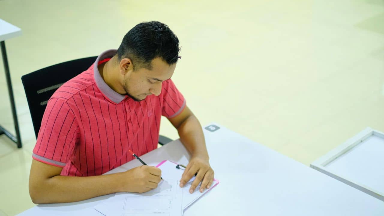 A man in a striped polo shirt is seated at a table, writing on a document.