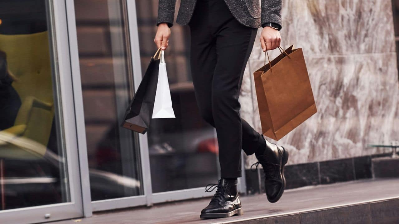 A person wearing a gray blazer and black pants is walking while holding two shopping bags.