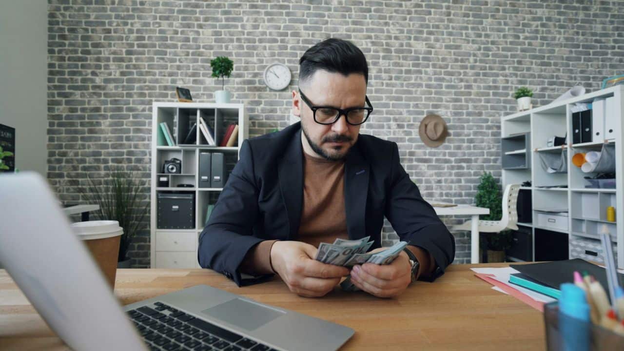 A man with a beard and glasses is sitting at a desk and counting money.