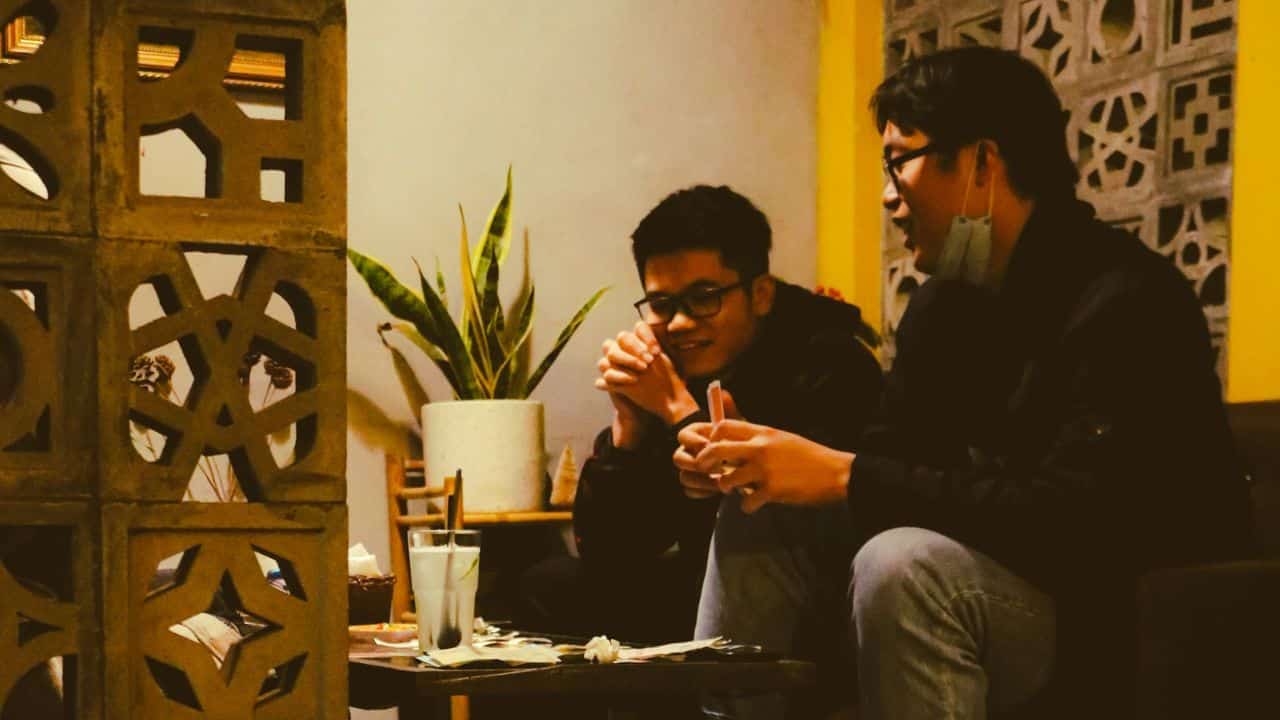 Two young men sitting at a table with drinks and talking in a dimly lit room.