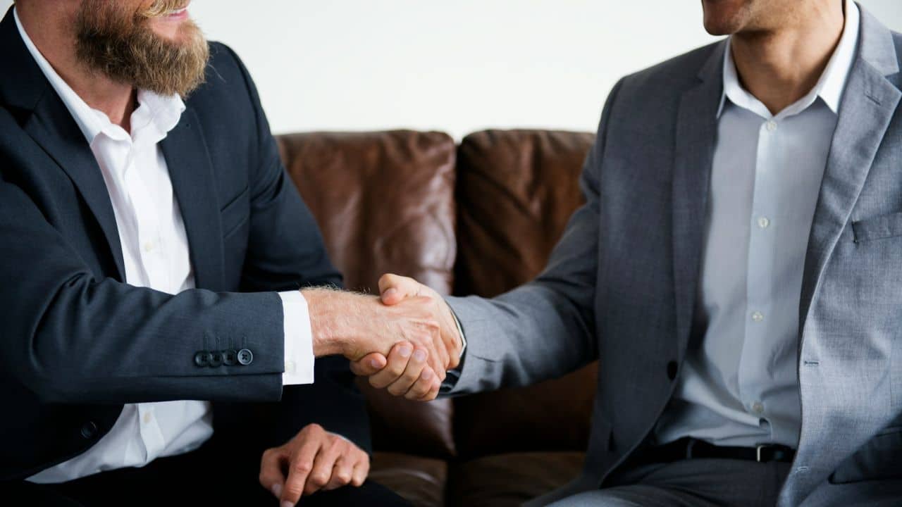 Two men in business suits are shaking hands over a brown couch.