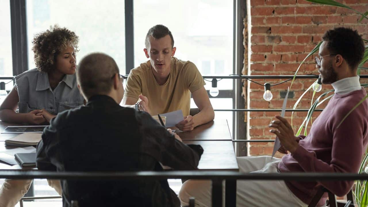 Four people are seated around a table, engaged in a conversation in a room with a brick wall.