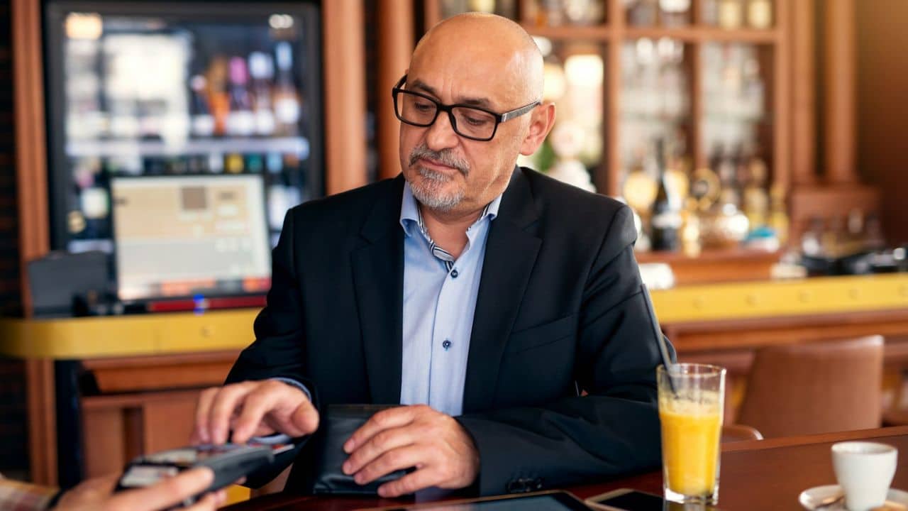 A bald man in glasses and a suit is paying with a credit card at a bar.