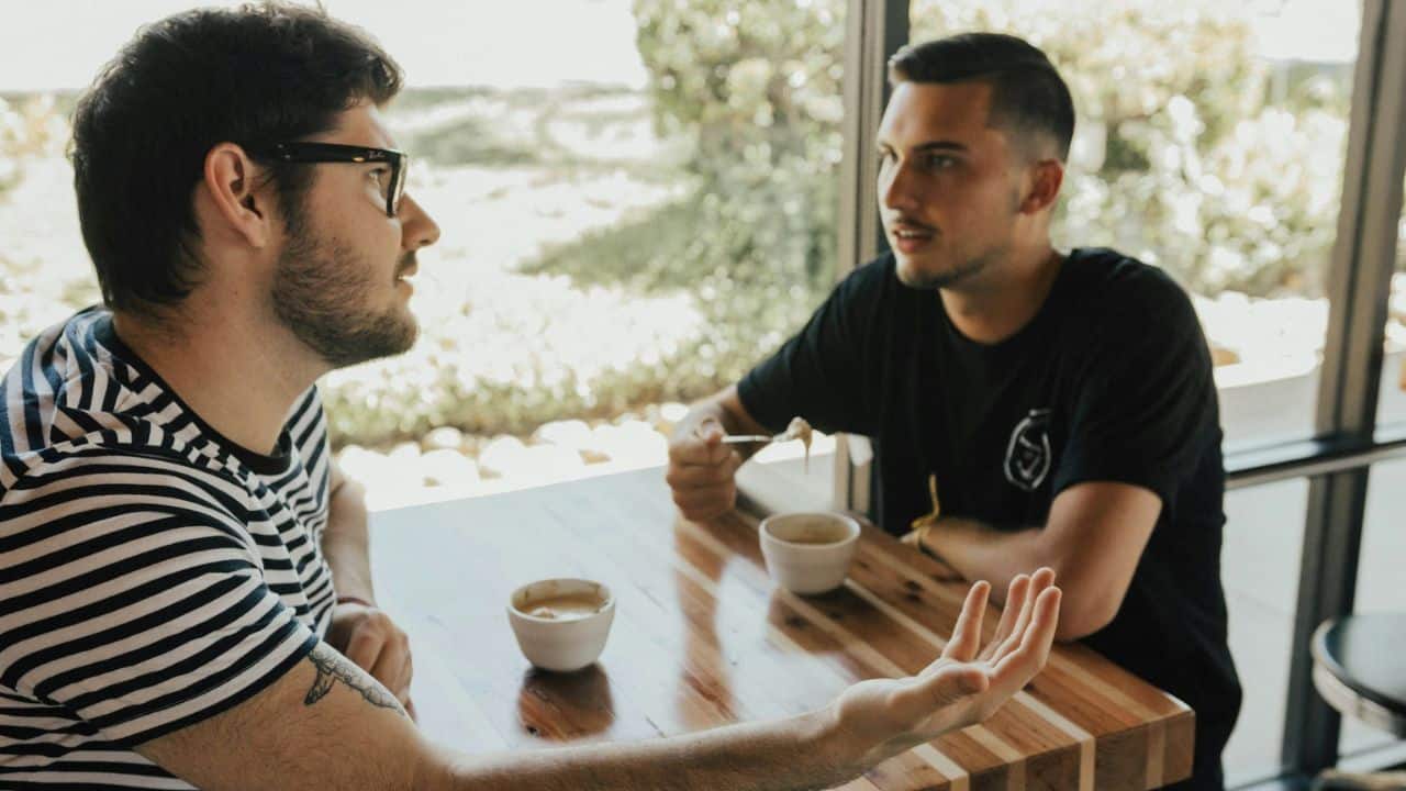 Two men are seated at a wooden table in a cafe, talking and gesturing.