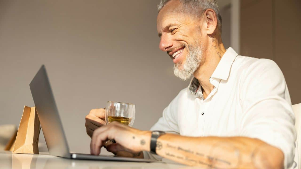 A gray-haired man with a beard is smiling while using a laptop and holding a glass.